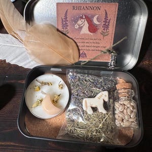 May include: An open metal tin containing a variety of items. Inside, there's a candle, a moon-shaped stone, dried herbs, a small horse figurine, and a glass jar with seeds. A feather and a card with the text "RHIANNON" are also present.