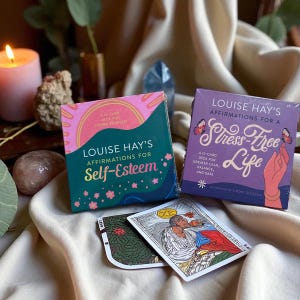 May include: Two card decks titled "Louise Hay's Affirmations for Self-Esteem" and "Louise Hay's Affirmations for a Stress-Free Life" are displayed. The decks are in boxes with colorful designs and text. Several affirmation cards are visible in front of the boxes.