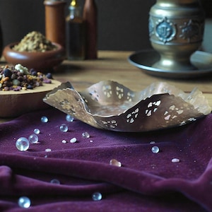 May include: A decorative, hammered metal bowl with a scalloped edge and cut-out floral designs. The bowl sits on a deep purple velvet cloth, with scattered clear glass beads. Other items in the background include wooden bowls and bottles.