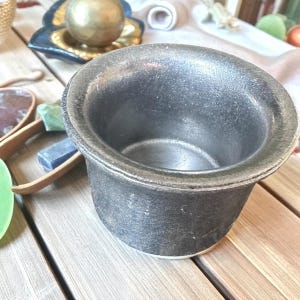 May include: A dark gray, textured ceramic bowl with a wide rim and a white base. The bowl is sitting on a wooden surface. Other objects, including a gold sphere, are visible in the background.