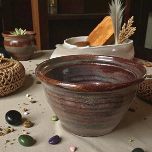 May include: A handmade, glazed ceramic bowl with a wide rim and a rich, earthy color palette of brown, rust, and hints of blue. The bowl is part of a collection of decorative pottery, perfect for home decor.