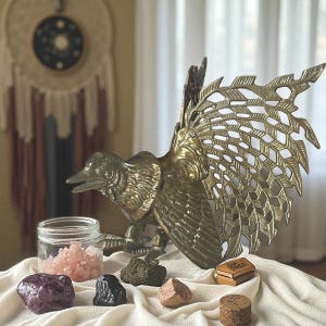 May include: A brass bird sculpture with openwork wings is the focal point of this image. Various crystals and small decorative items are arranged in front of the bird. A dream catcher hangs in the background, adding to the bohemian aesthetic.