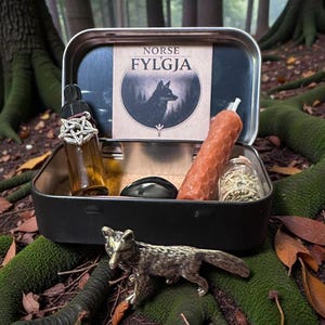May include: A black metal box with a "Norse Fylgja" label, containing a bottle, candle, and other items. A silver fox figurine sits in front of the box. The items are arranged on a mossy forest floor.