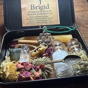 Puede incluir: Una caja de metal negro que contiene un kit de altar de Brigid. El kit incluye una tarjeta con texto, cristales, flores secas, velas, peque&ntilde;os frascos de vidrio y una cuchara decorativa. El texto de la tarjeta dice "Brigid, Diosa del Hogar, la Curaci&oacute;n y la Inspiraci&oacute;n."