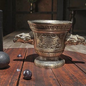 May include: A bronze-colored mortar and pestle with dragon head handles and ornate carvings. The mortar features a detailed relief of an animal, possibly a boar, and is set on a wooden surface. Decorative spheres and crystals are also present.
