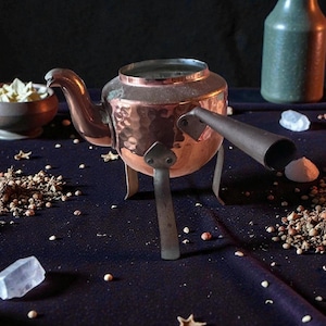 May include: A copper-colored teapot with a hammered texture, featuring a spout and a handle. The teapot is supported by three legs. A dark blue surface is scattered with small white objects and spices. A green bottle and a small bowl are in the background.