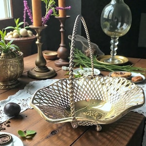 May include: A gold-colored, openwork metal basket with a twisted handle. The basket has a scalloped edge and sits on small feet. The background includes candles, a decorative vase, and other vintage items.