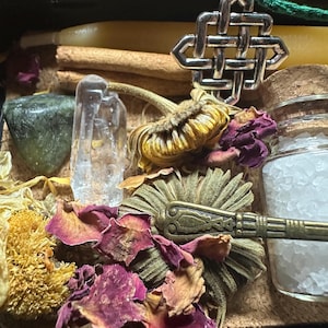 Puede incluir: Una colecci&oacute;n de art&iacute;culos espirituales que incluye un colgante de nudo celta plateado, p&eacute;talos de rosa secos, un cristal de cuarzo transparente, un peque&ntilde;o frasco de sal y una flor seca. Los art&iacute;culos est&aacute;n dispuestos en una caja negra.