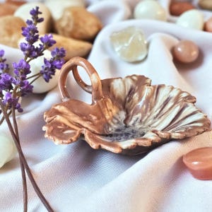 May include: A small, decorative ceramic dish in the shape of a leaf, featuring a brown and cream marbled design. The dish has a curved handle and is surrounded by lavender sprigs and small stones. The overall color palette is soft and earthy.