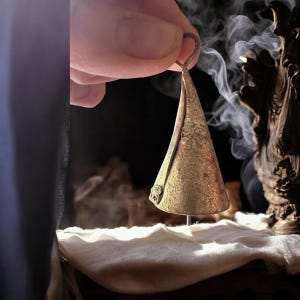 May include: A small, antique-looking brass bell with a conical shape. The bell has a loop at the top for hanging and a textured surface. The bell is held in a hand, with smoke in the background.