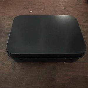 Puede incluir: Una caja rectangular de metal negro con esquinas redondeadas. La caja est&aacute; cerrada y descansa sobre una superficie de madera marr&oacute;n oscuro. La tapa tiene una superficie ligeramente reflectante.