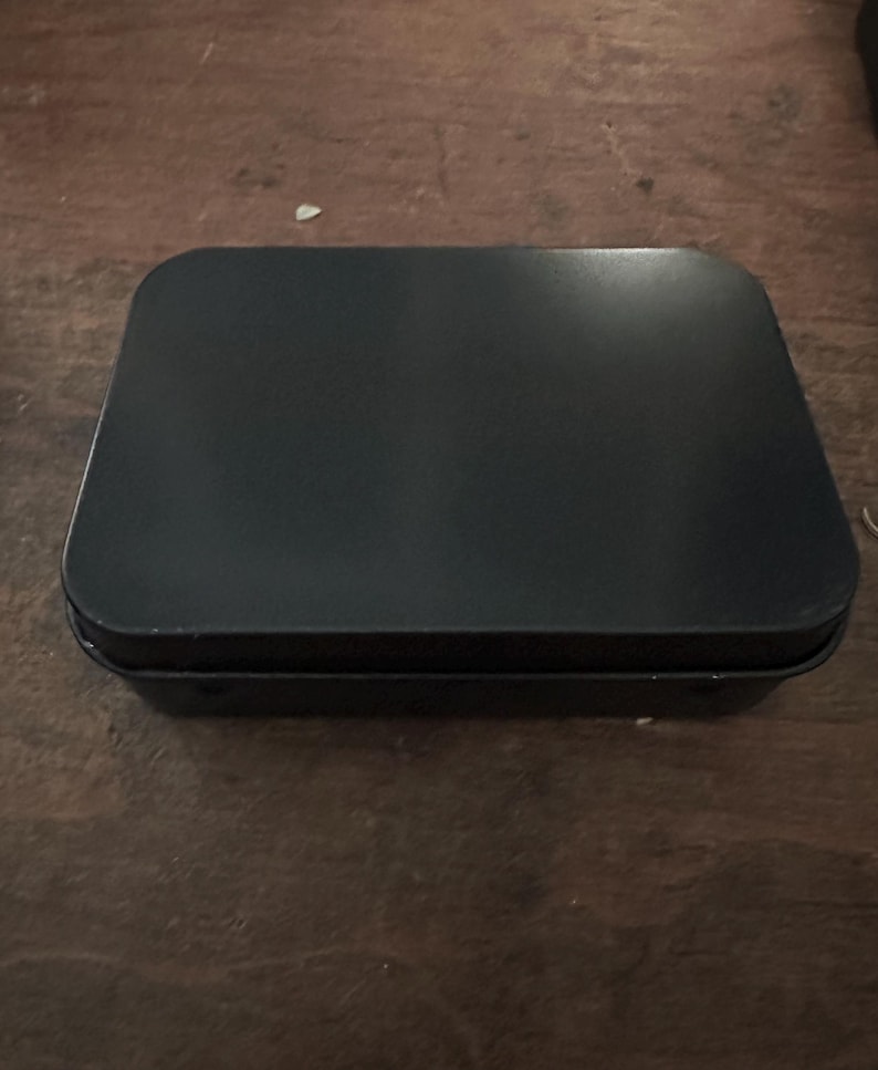 Puede incluir: Una caja rectangular de metal negro con esquinas redondeadas. La caja est&aacute; cerrada y descansa sobre una superficie de madera marr&oacute;n oscuro. La tapa tiene una superficie ligeramente reflectante.