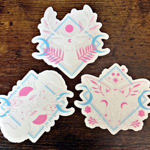 May include: Three white stickers with celestial designs. Each sticker features a central animal or insect motif, surrounded by geometric shapes, crescent moons, and pink and blue accents. The designs include a deer, a scorpion, and a moth.