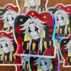 May include: Holographic sticker featuring a cartoon jester character with white hair, a sinister grin, and a red, black, and yellow harlequin outfit. The sticker has a star-shaped design on the top and is surrounded by other similar stickers.