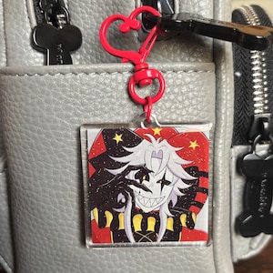 May include: A clear acrylic charm featuring a cartoon character with white hair, a black and white face, and a sinister grin. The charm is attached to a red heart-shaped clasp and a silver zipper. The background is a gray leather-like material.