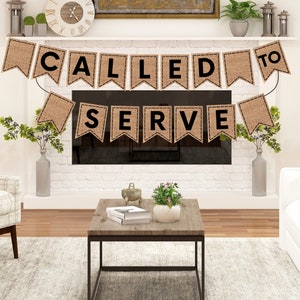Op de afbeelding: Een jute banner met de tekst "Called to Serve" in zwarte letters. De banner hangt boven een open haard in een woonkamer.