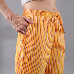 May include: Yellow and white striped trousers featuring thin vertical red lines, an elasticated waistband with a matching fabric drawstring, and side pockets. The garment is paired with a plain white short-sleeved top tied at the midriff.