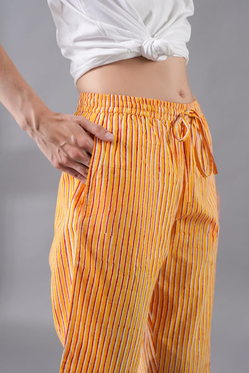 May include: Yellow and white striped trousers featuring thin vertical red lines, an elasticated waistband with a matching fabric drawstring, and side pockets. The garment is paired with a plain white short-sleeved top tied at the midriff.