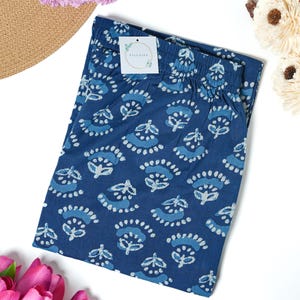 May include: Folded navy blue trousers with a white floral pattern. The trousers have an elasticated waistband and a tag that reads "Zillajee". The trousers are surrounded by pink tulips, a straw hat, and white decorative flowers.