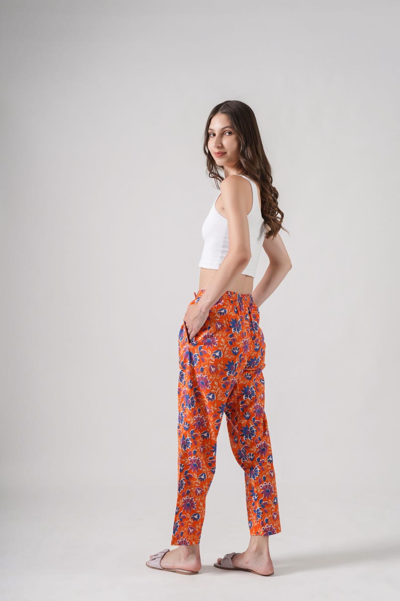 Handmade cotton pajama bottoms featuring colorful blue and purple flowers on bright orange fabric – soft, comfy, and breathable sleep pants designed with elastic waist, drawstring closure, and spacious side pockets, ideal for floral print clothing lovers, gifts for flower lovers, book lovers, and stylish cotton gift ideas.