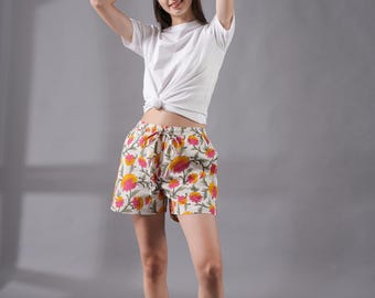 May include: A white t-shirt with a knotted front paired with floral print shorts. The shorts feature a cream background with pink and yellow flower patterns. The outfit is casual and summery.