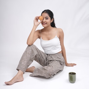 May include: A woman wearing a white camisole top and gray patterned pants, sitting on the floor. She holds a pastry to her eye. A green ceramic cup sits nearby. The pants have a drawstring waist and a leaf design.