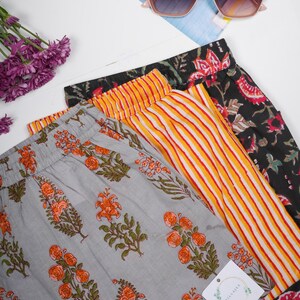 May include: Three pairs of trousers with elastic waistbands. One pair is light grey with an orange and green floral print. Another pair features vertical yellow and white stripes. The third pair is black with a red and green floral pattern.