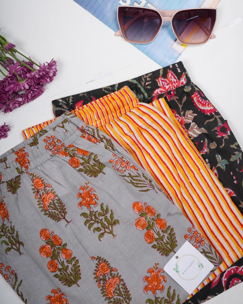 May include: Three pairs of trousers with elastic waistbands. One pair is light grey with an orange and green floral print. Another pair features vertical yellow and white stripes. The third pair is black with a red and green floral pattern.