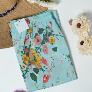 May include: Light blue floral print shorts with an elastic waistband. The shorts feature a colorful pattern of red, yellow, and pink flowers with green leaves. A straw hat and flowers are also in the image.