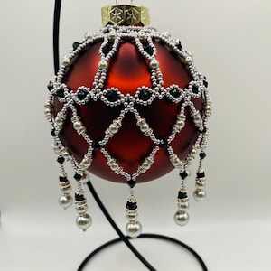 Peut inclure: Une boule de Noël en verre rouge avec un motif perlé noir et argent. La boule a une frange perlée et est exposée sur un support noir.