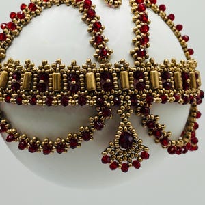 May include: A white spherical ornament adorned with a decorative beaded overlay. The intricate design features metallic gold seed beads and cylindrical beads combined with small, faceted deep red crystals arranged in a structured, crown-like pattern.
