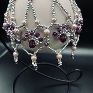 May include: An ornate bauble with a pale pink sphere, embellished with silver beads and purple and pink crystal accents. The ornament is displayed on a black metal stand, highlighting its detailed design. The beaded pattern gives a delicate, elegant look.