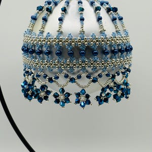 Pode incluir: Uma bola de Natal prateada com intrincados trabalhos de contas em tons de azul e prata. O ornamento apresenta um design de contas com detalhes florais pendurados, criando uma aparência festiva e elegante. É suspenso por uma tampa prateada.