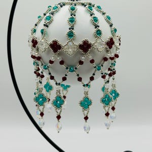 Puede incluir: Un adorno navideño blanco con intrincados abalorios en color turquesa, burdeos y cristal transparente. El diseño de abalorios incluye un patrón de enrejado y detalles colgantes de cristal, creando un aspecto festivo y elegante.