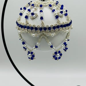 Peut inclure: Une boule de Noël blanche ornée de perles complexes. L'ornement présente un motif de perles bleues et argentées, avec des accents de perles. Le design comprend des pampilles perlées et des grappes florales, créant une apparence festive et élégante.