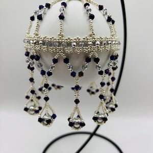 May include: A white Christmas ornament with silver and dark blue bead accents. The ornament has a beaded fringe with silver beads and dark blue crystal beads. The ornament is hanging from a silver metal cap and a black metal stand.