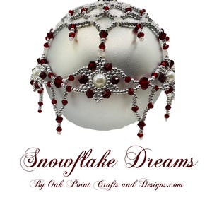 以下が含まれることがあります： 赤と銀のビーズで雪の結晶模様が施された銀色のクリスマスオーナメント。オーナメントは赤いビーズと白い真珠で飾られています。オーナメントの下には「Snowflake Dreams By Oak Point Crafts and Designs.com」という文字が書かれています。