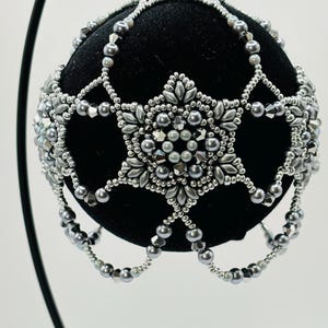 May include: A spherical black ornament decorated with a complex, hand-beaded silver overlay. The design features a central star-like pattern composed of metallic gray seed beads, faceted crystals, and smooth pearls, creating a draped, net-like effect.
