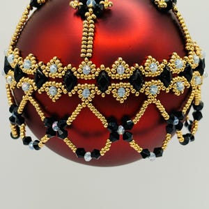 Puede incluir: Un adorno esférico rojo decorado con una red intrincada de cuentas doradas, cuentas negras en forma de diamante y pequeños cristales iridiscentes. El trabajo de cuentas forma un patrón de celosía estructurado sobre la esfera roja.