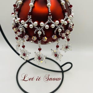 May include: A red glass bauble with a white and silver beaded design and snowflake accents. The bauble is on a black stand and the text "Let it Snow" is written below the bauble.