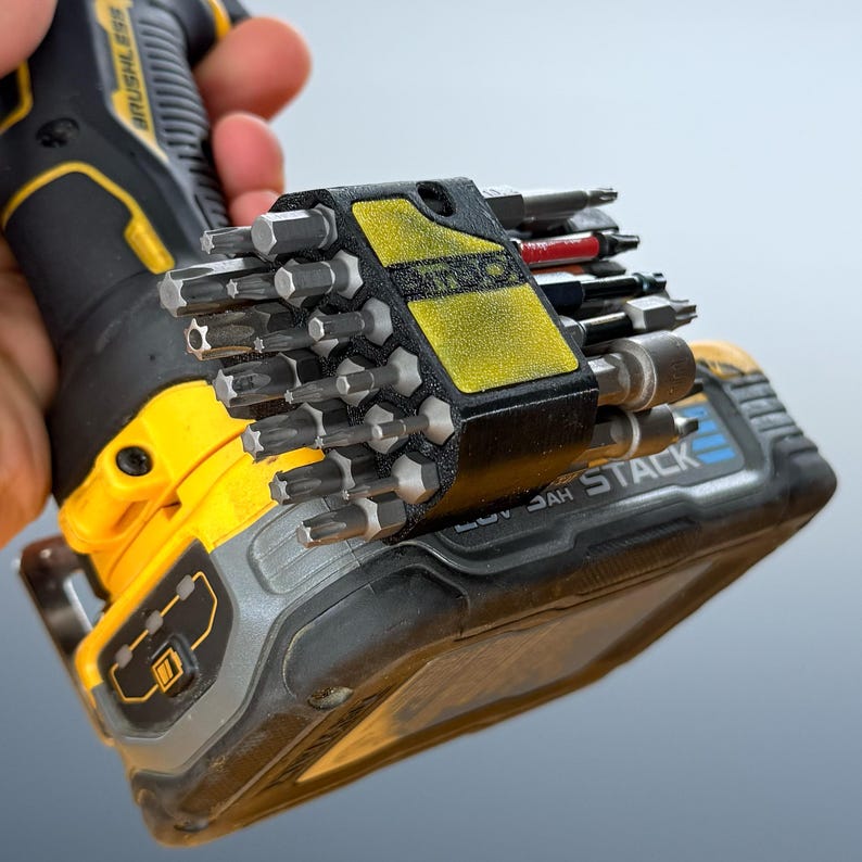 May include: A black and yellow power drill with a 20V battery pack attached. A black 3D-printed bit holder is mounted to the side of the battery, containing a variety of metal screwdriver bits with star and hexagonal heads in different sizes.