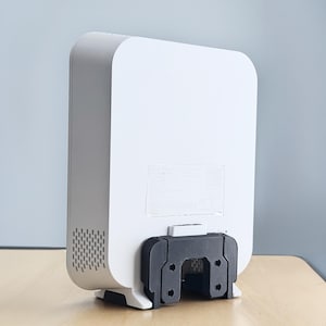 May include: A white rectangular device with rounded edges, featuring a black mounting bracket on the bottom. The device has a vent on the side and a sticker on the back. It is placed on a wooden surface.