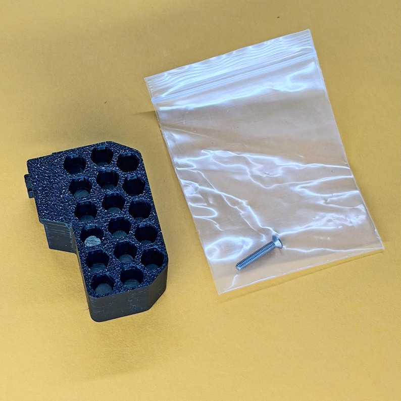 May include: A black, textured, 3D-printed component featuring a grid of twelve hexagonal openings arranged in a block shape, accompanied by a single silver-coloured metal screw enclosed in a small, clear, rectangular plastic zip-top bag.