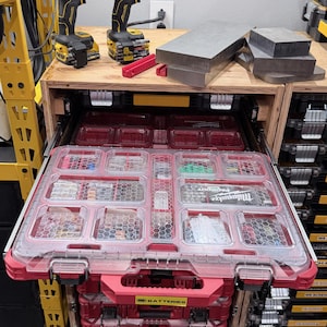 K&ouml;nnte beinhalten: Ein rotes und transparentes Milwaukee Packout Schubladensystem mit mehreren F&auml;chern f&uuml;r verschiedene Kleinteile. Zwei gelb-schwarze Akku-Bohrschrauber stehen auf der h&ouml;lzernen Werkbank &uuml;ber den gestapelten roten Aufbewahrungseinheiten.