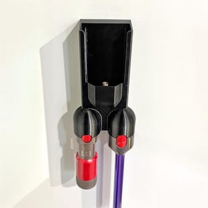 Könnte beinhalten: Ein schwarzer, an der Wand montierter Zubehörhalter für Staubsauger. Der Halter hat zwei Andockports mit roten Knöpfen und ein Ablagefach. Ein roter und silberner Staubsaugeraufsatz und ein lila Stab sind angedockt.