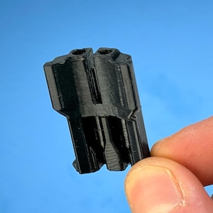 May include: A black, 3D-printed object with a unique, angular design. The object has two cylindrical openings at the top and a series of ridges and angles along its sides. The item is held against a blue background.