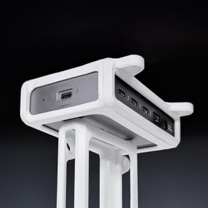 May include: A white plastic docking station mount holding a rectangular electronic hub. The hub features a grey front panel with a USB-C port labeled Computer, and a side panel with multiple ports including USB-C, USB-A, and a circular power input.