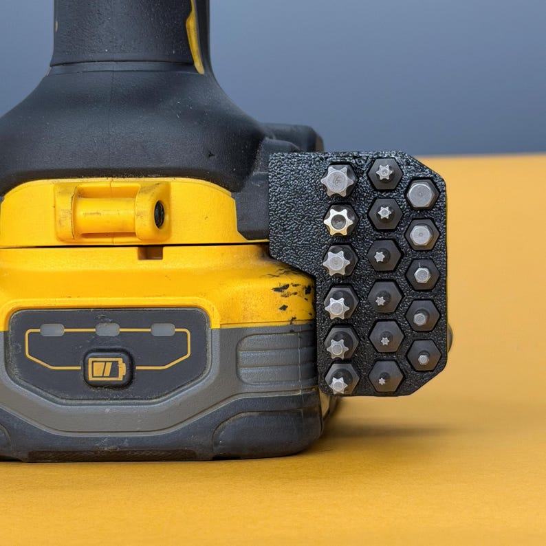 May include: A black and yellow power tool battery base featuring a black, textured, multi-slot bit holder attached to the side. The holder contains various metal screwdriver bits, including star and hexagonal shapes, arranged in a grid pattern.