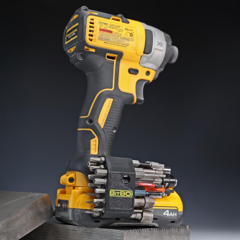May include: A yellow and black cordless impact driver with a brushless motor and a 4Ah battery. Attached to the battery base is a black BitBoi bit holder containing a variety of metal screwdriver bits and sockets for professional power tool use.
