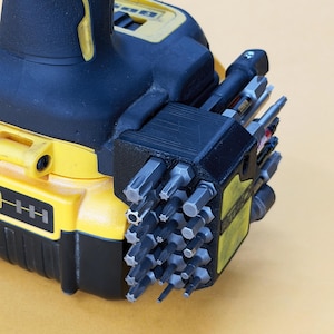 May include: A black 3D-printed bit holder attachment mounted to a yellow and black power drill. The holder contains a grid of various metal screwdriver bits, including star, hex, and flathead tips, arranged in rows for organised storage.